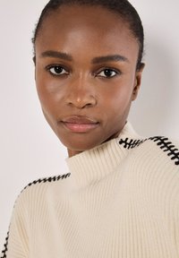 Cremfarbener, gerippter Pullover mit hohem Kragen und schwarzen Nähakzenten an den Schultern. Glatte Textur mit tailliertem Schnitt.