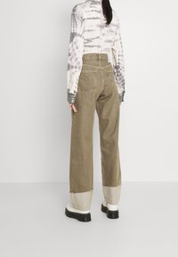 Pantalones de pierna ancha en verde oliva con puños beige en contraste, fabricados en un tejido estructurado. Los bolsillos traseros son visibles con una etiqueta de la marca.