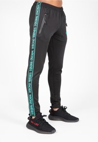 Zwarte joggingbroek met elastische tailleband, zijritsen en turquoise logo op de buitennaad. Gecombineerd met zwarte sneakers.
