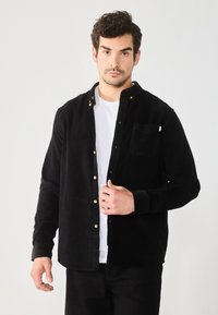Black corduroy shirt with gold buttons, button-down collar, single chest pocket, long sleeves, and textured fabric, layered over a white t-shirt.