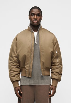 Homme portant une veste aviateur beige sur un pull en maille beige et un pantalon marron, debout devant un fond clair uni.