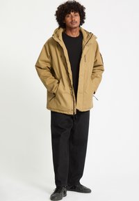 Khaki pufferjakke med hætte, der har en frontlynlås, snøre og logodetalje. Sammen med sorte loose-fit bukser og sorte sko.