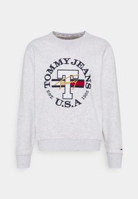 Šedá mikina s kulatým výstřihem, která má velké námořnické modré a červené kruhové logo obsahující tučný text "TOMMY JEANS U.S.A." a zlaté akcenty.