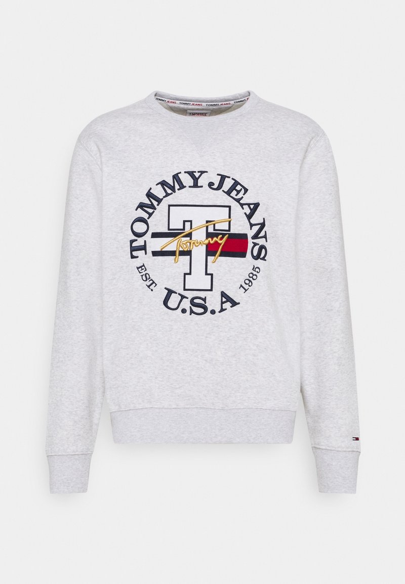Šedá mikina s kulatým výstřihem, která má velké námořnické modré a červené kruhové logo obsahující tučný text "TOMMY JEANS U.S.A." a zlaté akcenty.