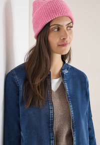 Veste en jean bleu foncé avec fermeture éclair, portée sur un pull léger. Bonnet en tricot côtelé rose sur la tête. Texture et formes subtiles.