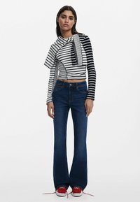 Gestreiftes schwarz-weißes Oberteil mit asymmetrischem Design, kombiniert mit dunkelblauen Schlagjeans. Rote Schuhe mit weißen Akzenten vervollständigen das Outfit.