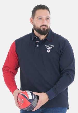 Homme avec une barbe tenant un ballon de rugby rouge et marine, portant une chemise marine avec une manche gauche rouge et le logo "Ruckfield" sur la poitrine.