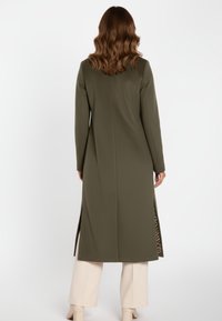 Lange olijfgroene jas met een gestructureerde afwerking, voorzien van zijopeningen en een luipaardprintaccent. Gedragen over lichtgekleurde broek.