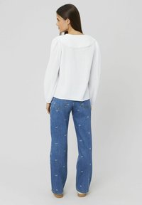 Blouse blanche texturée avec un col festonné, associée à un jean en denim bleu ornés de motifs brodés de petits nœuds blancs.
