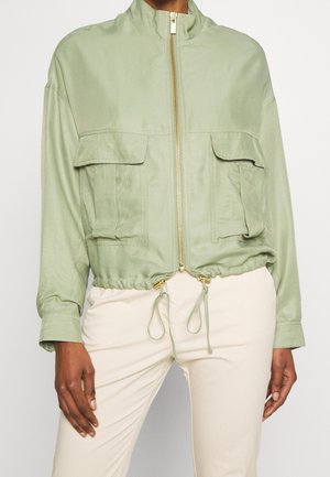 Chaqueta de color verde claro con cremallera, bolsillos delanteros con solapa y dobladillo ajustable con cordón, llevada con pantalones crema.
