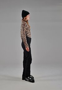 Ung kvinde i sort hue, leopardprint top, sorte bukser og skistøvler står sideværts mod en grå baggrund.