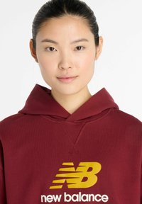Maroon hoodie with a large yellow "new balance" logo. Features a drawstring hood and ribbed hem, with a soft texture.