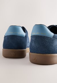 Blå ruskindssneakers med en glat blå læderhæl og en brun gummiydersål, med prægede tekster og et krone-logo på hælen.