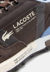 Καφέ αθλητικό παπούτσι από ύφασμα και σουέτ με μπλε λεπτομέρειες. Διαθέτει λευκή σόλα, σχέδιο με λογότυπο και συντεταγμένες τυπωμένες στην πλευρά.