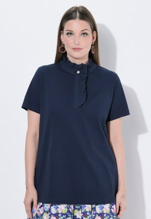 Donna che indossa una maglietta blu navy a maniche corte con collo alto arricciato e gonna floreale, in piedi davanti a uno sfondo bianco semplice.