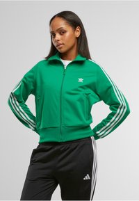 Chaqueta verde con cremallera fabricada en tela suave, con detalles de tres franjas blancas en las mangas y un logo bordado en el pecho.