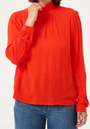 Kobieta ubrana w jasnoczerwoną bluzkę z długim rękawem z marszczonym dekoltem i mankietami, w połączeniu z niebieskimi dżinsami, z ręką przy twarzy.