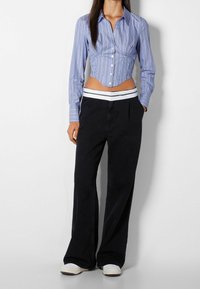 Blå och vit randig kortärmad skjorta med långa ärmar, frampanelknappar och samlade sidor, tillsammans med vida svarta byxor och vita sneakers.