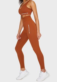 Femme portant un brassière de sport côtelée de couleur rouille et un legging taille haute avec le logo Squatproof, chaussée de baskets blanches, debout de profil.