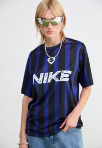 Camiseta de Nike con rayas verticales azules y negras, gran logo blanco, mangas cortas y un corte relajado, acompañada de gafas de sol metálicas.