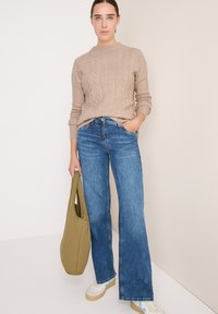 Beige Kabelstrickpullover, blaue Schlagjeans, beige Sneakers mit weißer Sohle, hält eine olivgrüne Tasche. Flache Textur, lässige Passform.