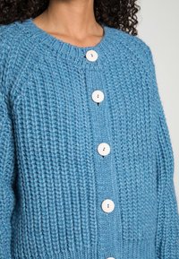 Pletený kardigan ve světle modré barvě s kulatým výstřihem, který má pět velkých bílých knoflíků a ribovaný vzor na předním panelu.