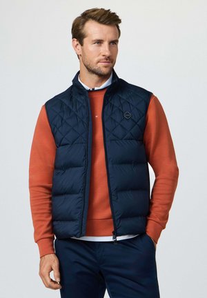 Man die een marineblauw doorgestikt vest draagt over een oranje trui en marineblauwe broek, staand met één hand in de zak tegen een effen achtergrond.