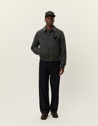 Grijze gestreepte bomberjack met kraag, zwart geborduurd logo, gecombineerd met donkere broek en een bijpassende gemustreerde pet. Bruine schoenen maken de look compleet.