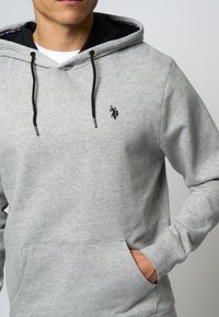 Šedá mikina s kapucí, černými šňůrkami, přední kapsou a malým černým vyšitým logem. Měkká látka s hladkým povrchem a uvolněným střihem.