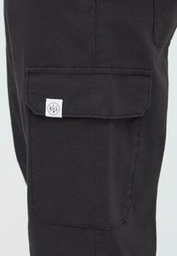 Crne cargo hlače imaju bočnu džepu s bijelom etiketom. Izrađene su od čvrstog materijala, posjeduju elegantan, minimalistički dizajn s čistim linijama.