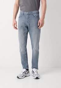 Jeans en denim bleu clair avec une coupe droite, design classique à cinq poches et un léger délavage. Portés avec des chaussures de sport grises et blanches.