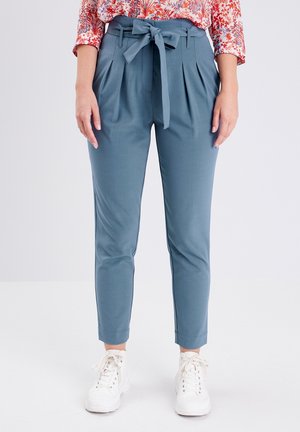 Femme portant un pantalon bleu taille haute avec ceinture nouée, un chemisier fleuri et des baskets blanches, debout devant un fond blanc uni.