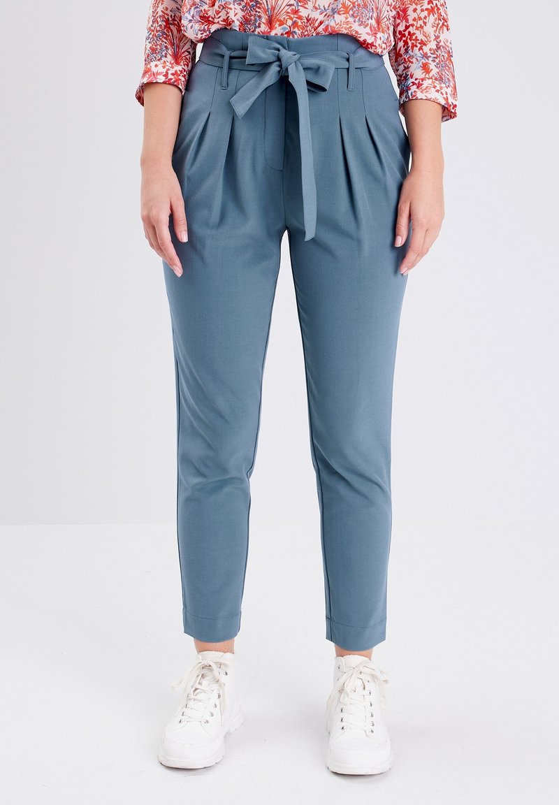 Femme portant un pantalon bleu taille haute avec ceinture nouée, un chemisier fleuri et des baskets blanches, debout devant un fond blanc uni.