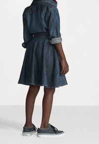 Jeansklänning med åtsittande liv och utsvängd kjol, med uppkavlade ärmar. Kombination med mörkgrå sneakers och en synlig logotyp.