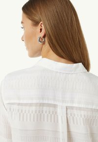 Weiße Bluse mit vertikalen Streifen, strukturierter Stoff, geknöpfter Kragen, Rückenfalte, akzentuiert durch einen dezenten Glanz. Sichtbare silberne Creolen.