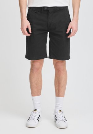 Männlich, trägt schwarze Shorts bis zum Knie, weiße Socken bis zur Mitte der Wade und weiße Adidas-Sneakers mit schwarzen Streifen auf einem einfarbigen Hintergrund.
