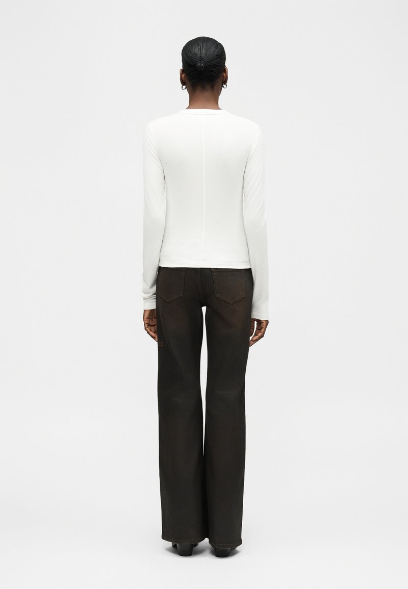 Femme avec les cheveux attachés en chignon, portant un haut blanc à manches longues et un pantalon noir à jambes larges, debout face à un fond blanc uni.