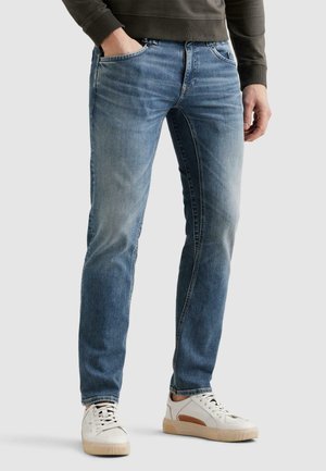 Mann trägt verblasste blaue Jeans mit Hand in der Tasche und weiße Sneakers mit beigen Sohlen, steht vor einfachem Hintergrund.