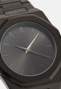 Montre octogonale avec un boîtier en métal noir mat, un cadran gris, des index en argent et des aiguilles fines, le tout accompagné d'un bracelet en métal texturé.