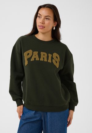 Femme aux longs cheveux foncés portant des boucles d'oreilles créoles dorées, un pull vert foncé avec le texte "PARIS" et un jean bleu, regardant légèrement sur le côté.