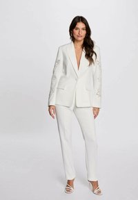 Blazer bianco con ricami floreali sulle maniche, abbinato a pantaloni su misura coordinati e décolleté bianchi con cinturini. Tessuto liscio e strutturato.