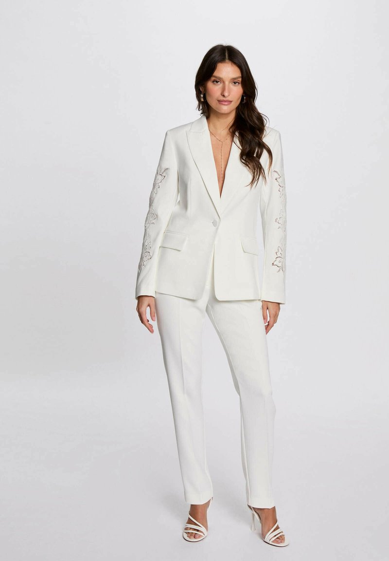 Blazer bianco con ricami floreali sulle maniche, abbinato a pantaloni su misura coordinati e décolleté bianchi con cinturini. Tessuto liscio e strutturato.