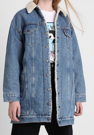 Lyseblå denimjakke med en krave af shearlingskind, store frontlommer, metalknapper og en afslappet pasform. Bæres over en T-shirt med grafik.