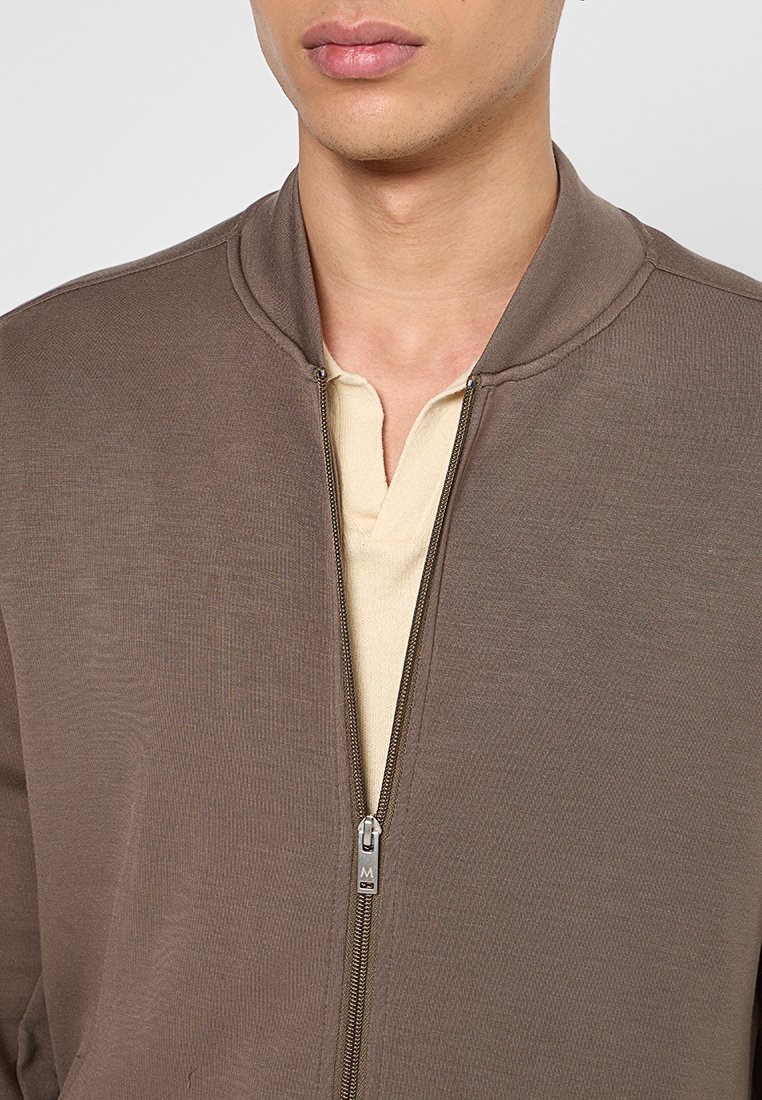 Brązowa kurtka na zamek z teksturą w prążki i okrągłym kołnierzem. Posiada kontrastową jasnobeżową koszulę z dekoltem na trzy guziki.