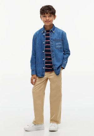 Jongen staat met een blauw denim overhemd over een gestreept marineblauw shirt, beige broek en witte sneakers tegen een effen lichte achtergrond.