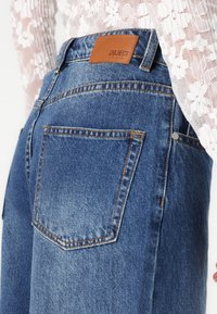 Jeans din denim albastru cu talie înaltă, având un eticheta din piele pe spate, cusături portocalii și două buzunare pe spate.