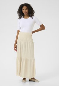 Maxi skirt in light beige fabric with a textured finish, featuring an elastic waistband and tiered design. Paired with a white short-sleeve top.