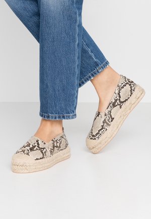 Personne portant un jean bleu et des chaussures beige à enfiler avec un imprimé serpent noir et marron et des semelles compensées tissées.