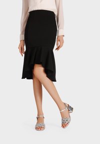 Gonna nera con orlo svasato, abbinata a sandali argentati con un motivo decorativo e un tacco basso, che presentano un aspetto raffinato.