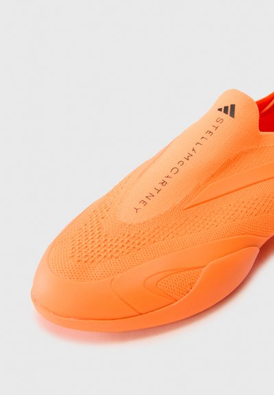 Oransje slip-on sneakers med et teksturert strikket overdel, gummisåle og subtil merkevare, med en strømlinjeformet form og glatt finish.